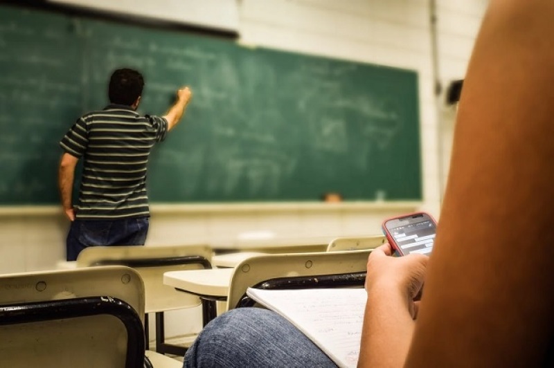 teacher writing on the classroom blackboard while a student tinkers with a phone